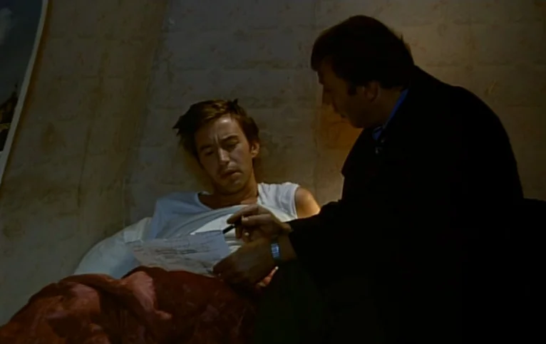 Giliap (1975) movie still: medium — A small room with light-colored textured wallpaper and a bed in the foreground. A man sitting on the side of the…
