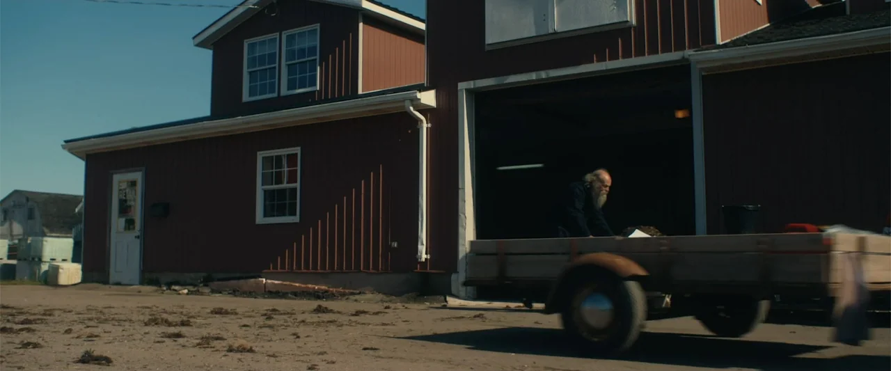 A Small Fortune (2021) movie still: wide — A red wooden barn with a large open bay sits on a dirt lot. An older man with a long beard works near a…