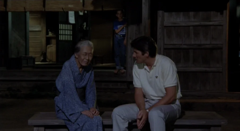 Rhapsody in August (1991) movie still: medium — An elderly Japanese woman and a Caucasian man sit together on a wooden bench. A young boy stands in…