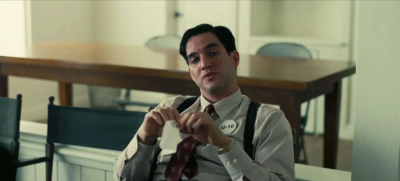 Oppenheimer (2023) movie still: medium — A spartan meeting room contains a long wooden table and simple chairs. A man in a collared shirt sits and…