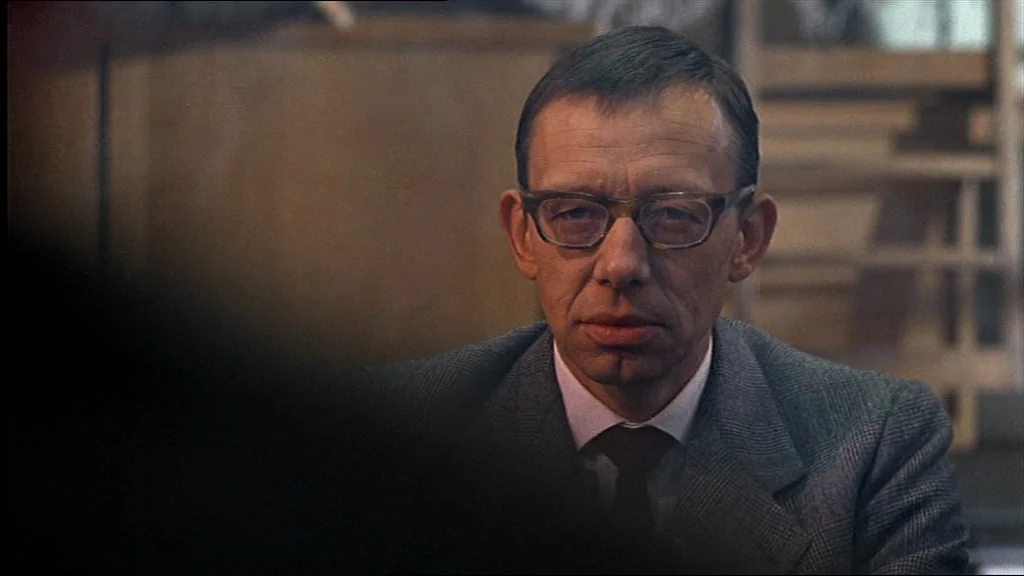 Lola (1981) movie still: closeup — A man in a grey suit sits in an indoor space with blurry background. He stares directly ahead at someone…