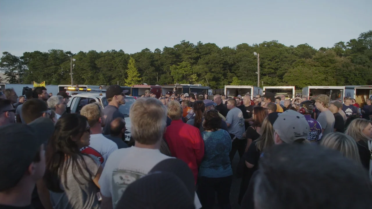 The Last Race (2022) movie still: wide — A dense crowd of people gathers in a parking area near several enclosed trailers. The group stands facing…