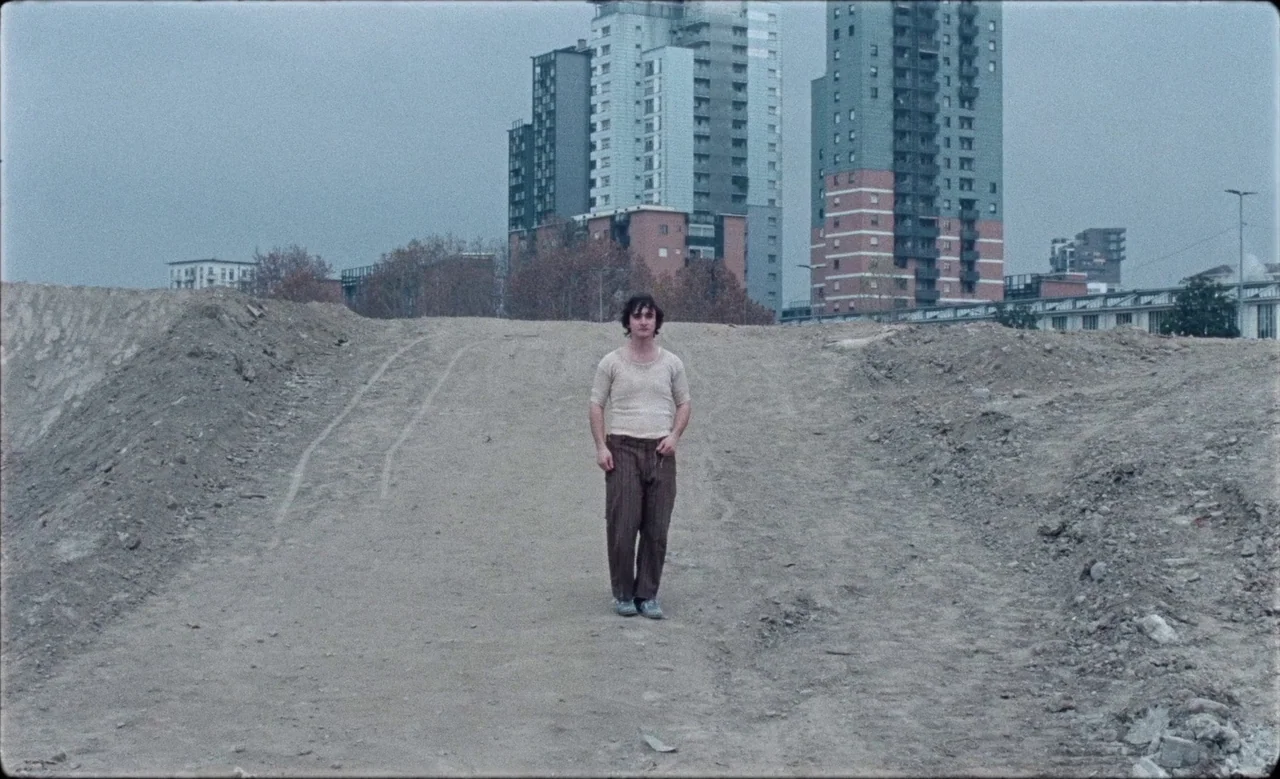 Happy as Lazzaro (2018) movie still: fullbody — An expanse of barren dirt mounds occupies a construction site under a gray sky. A lone man stands…