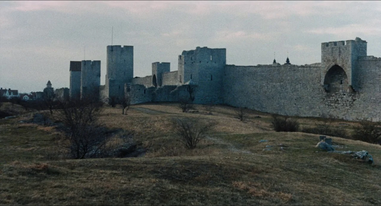 The Touch (1971) movie still: establishing — An extensive medieval stone wall with multiple fortified towers stretches across a grassy field. The…