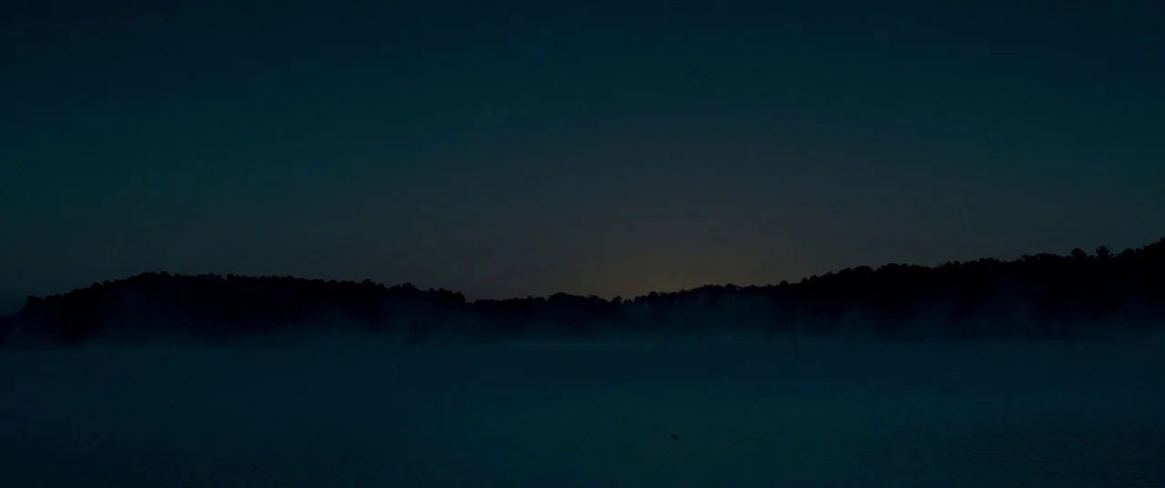 Lawless (2012) movie still: establishing — A vast lake surface is blanketed by a thick layer of morning mist. Dense treelines frame the dark horizon…