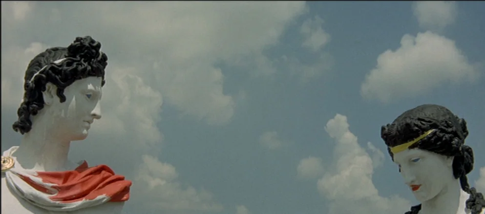 Contempt (Le Mépris) (1963) movie still: medium — Two classical-style stone busts are positioned against a bright blue sky with white clouds. The…