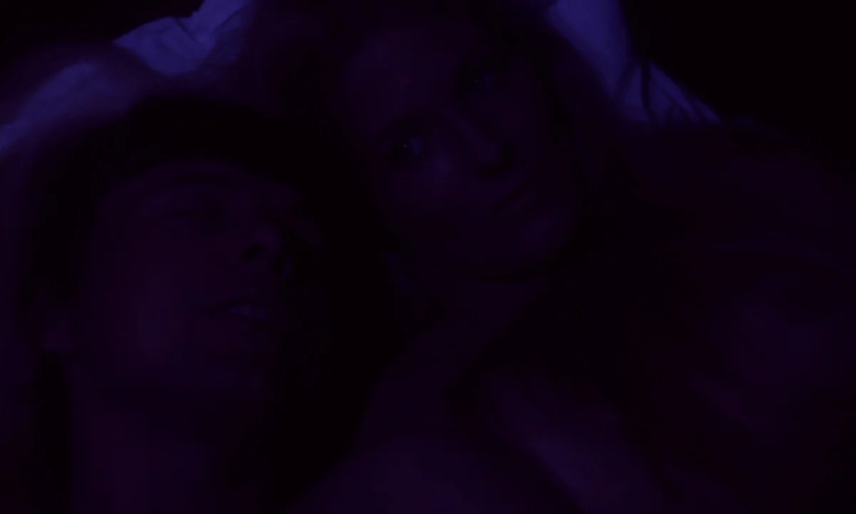 Daughters of Darkness (1971) movie still: closeup — A dark bedroom with two people lying on white pillows. The scene is illuminated by dim violet…