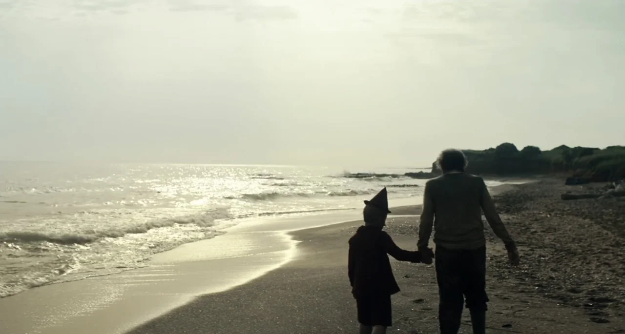 Pinocchio (2022) movie still: wide — A wide beach features gentle waves rolling onto the sand under a hazy overcast sky. An elderly man and a young…