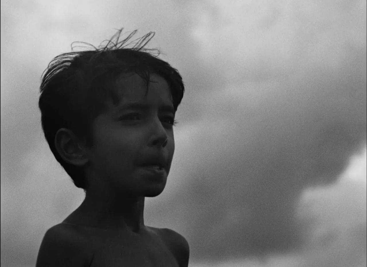 Pather Panchali (1955) movie still: closeup — A young boy stands in front of a wide, overcast sky filled with clouds. He gazes off-camera to the…