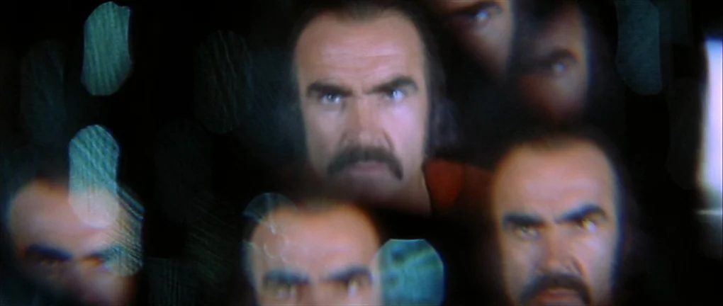 Zardoz (1974) movie still: closeup — A dark space is filled with multiple overlapping portraits of the same man. Each face stares directly forward…