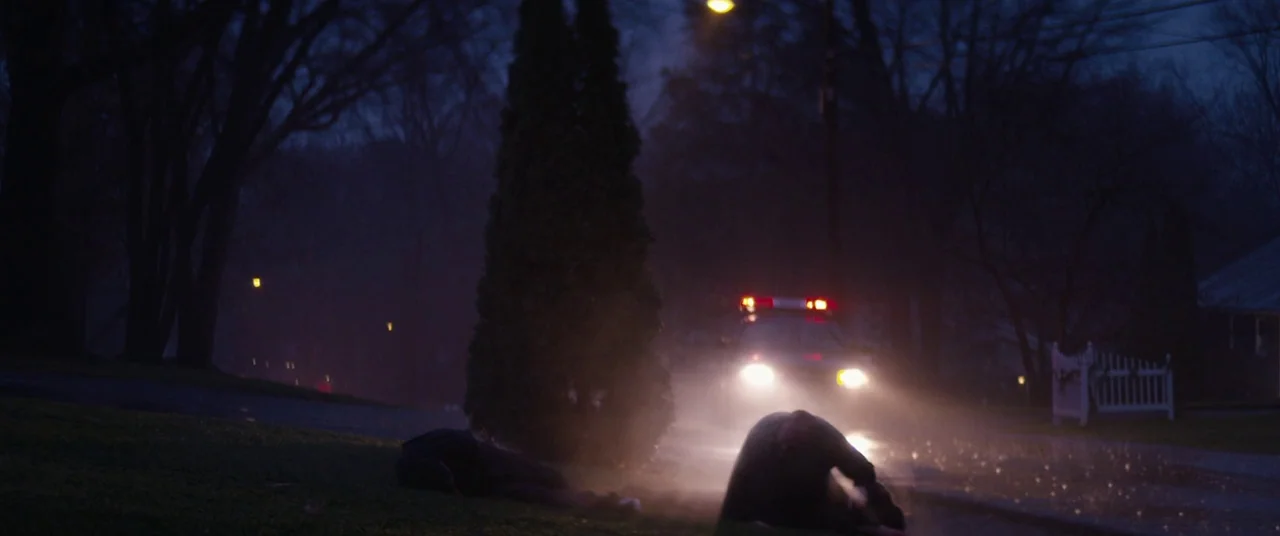 Super Dark Times (2017) movie still: wide — A dark, misty residential street at night featuring a stationary police car with flashing lights. Two…