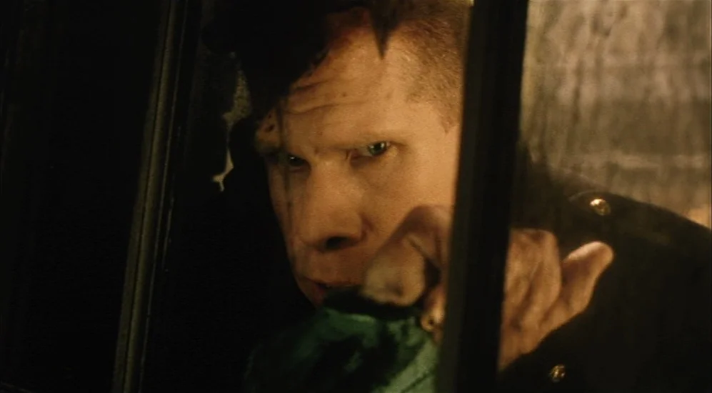 The City Of Lost Children movie still: closeup — A man peers through a dark pane of glass inside a vehicle interior. The scene is illuminated by…
