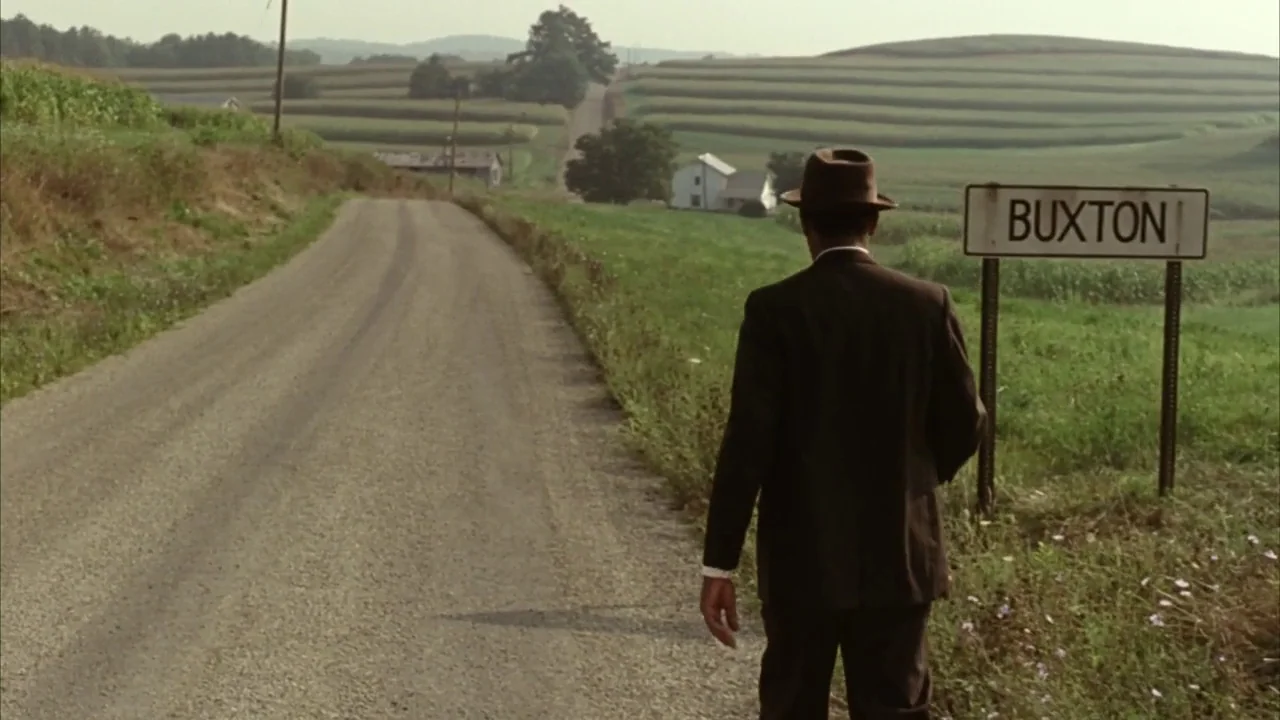 The Shawshank Redemption (1994) movie still: fullbody — A gravel country road cuts through rolling agricultural fields under a hazy sky. A man in a…