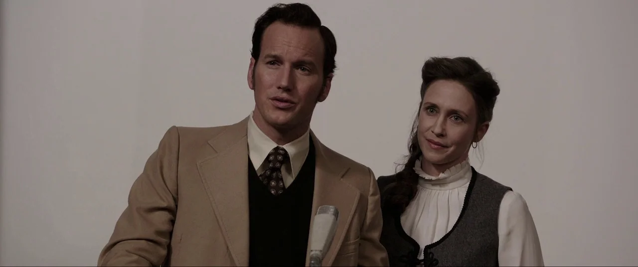 The Conjuring (2013) movie still: medium — A plain white background serves as the backdrop for a formal interview setting. A man in a tan blazer and…