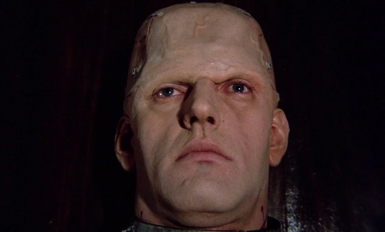 The Horror of Frankenstein (1970) movie still: closeup — The face of a man with prosthetic staples lies against a dark background. He gazes directly…