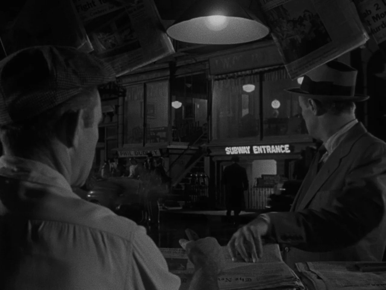 Pickup on South Street (1953) movie still: medium — A dark urban scene features a newsstand with newspapers hanging from an overhead structure. Two…