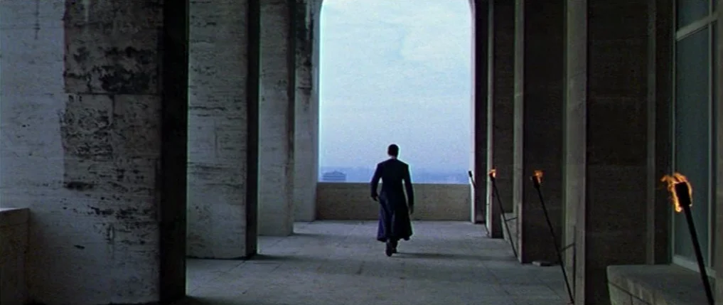 Titus (1999) movie still: wide — A stone balcony with large columns faces a bright, overcast outdoor horizon. A lone man in a dark coat walks away…