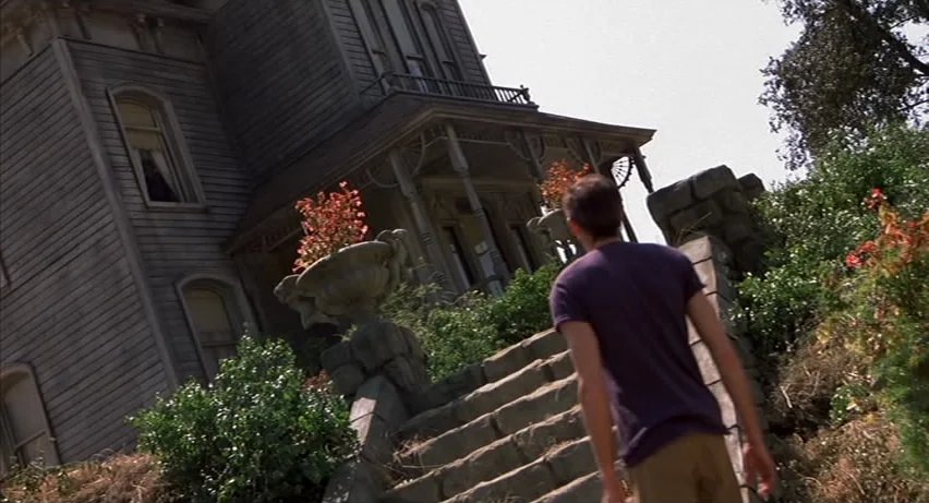 Psycho 2 (1983) movie still: wide — A large grey Victorian-style house stands behind stone steps with stone urn planters. A teenage boy in a purple…