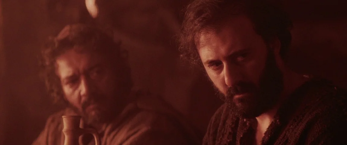 The Passion of the Christ (2004) movie still: medium — An interior space lit by firelight shows stone or earthen walls in the background. Two…