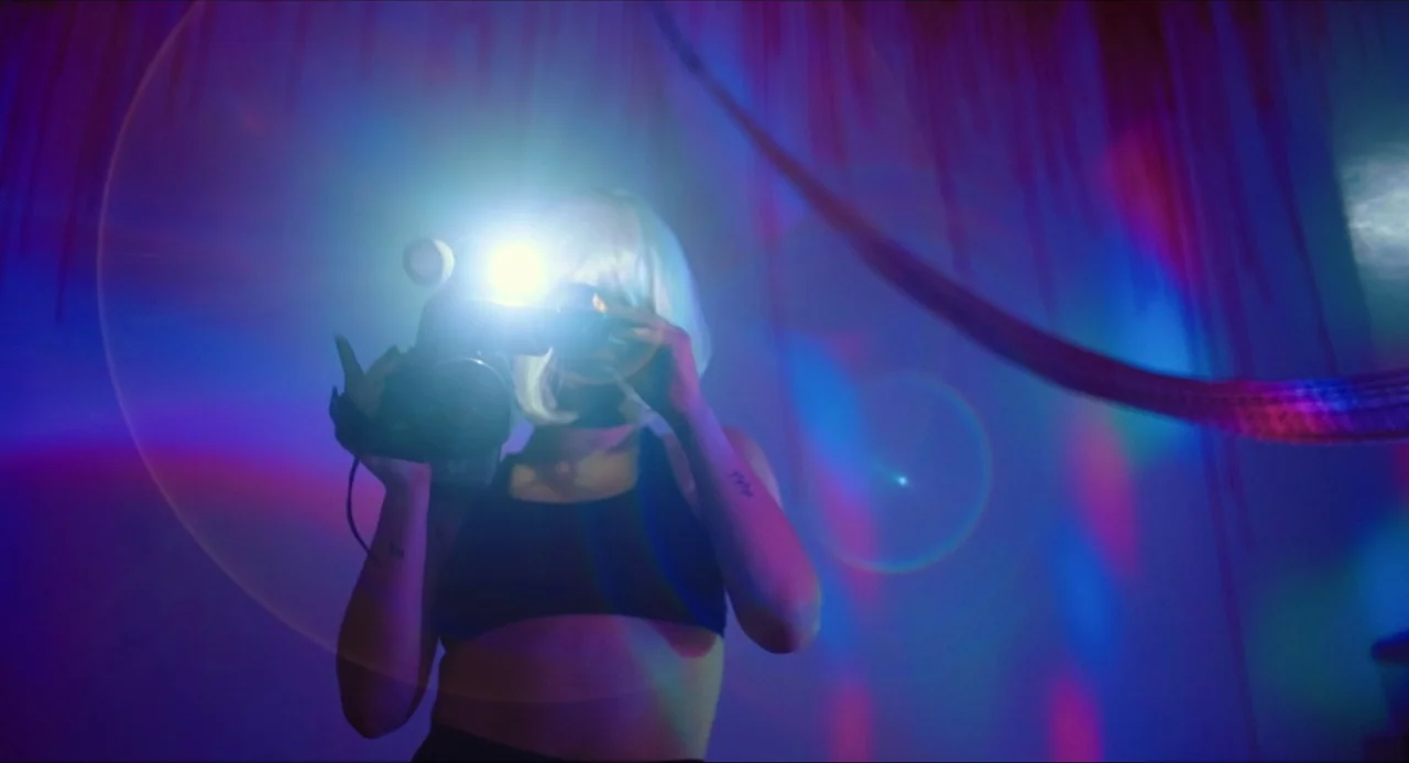Like Me movie still: medium — An interior space bathed in intense blue and magenta neon light beams. A person wearing a platinum blonde wig operates…