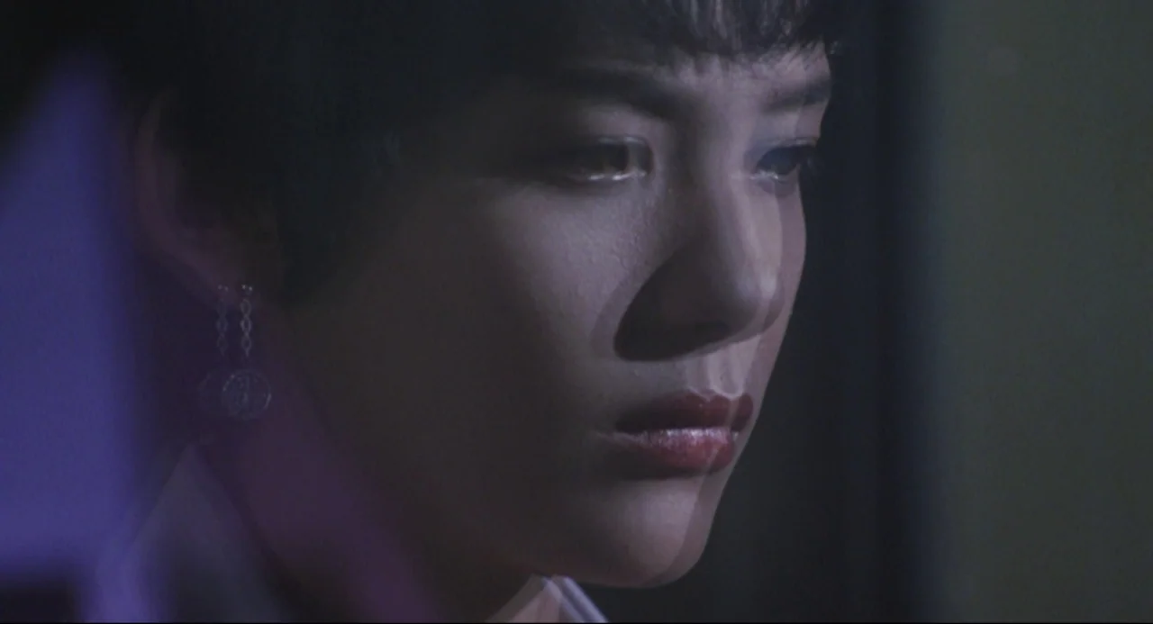 Godzilla vs. SpaceGodzilla (1994) movie still: closeup — A dimly lit indoor space with a neutral wall in the background. A woman with short dark…