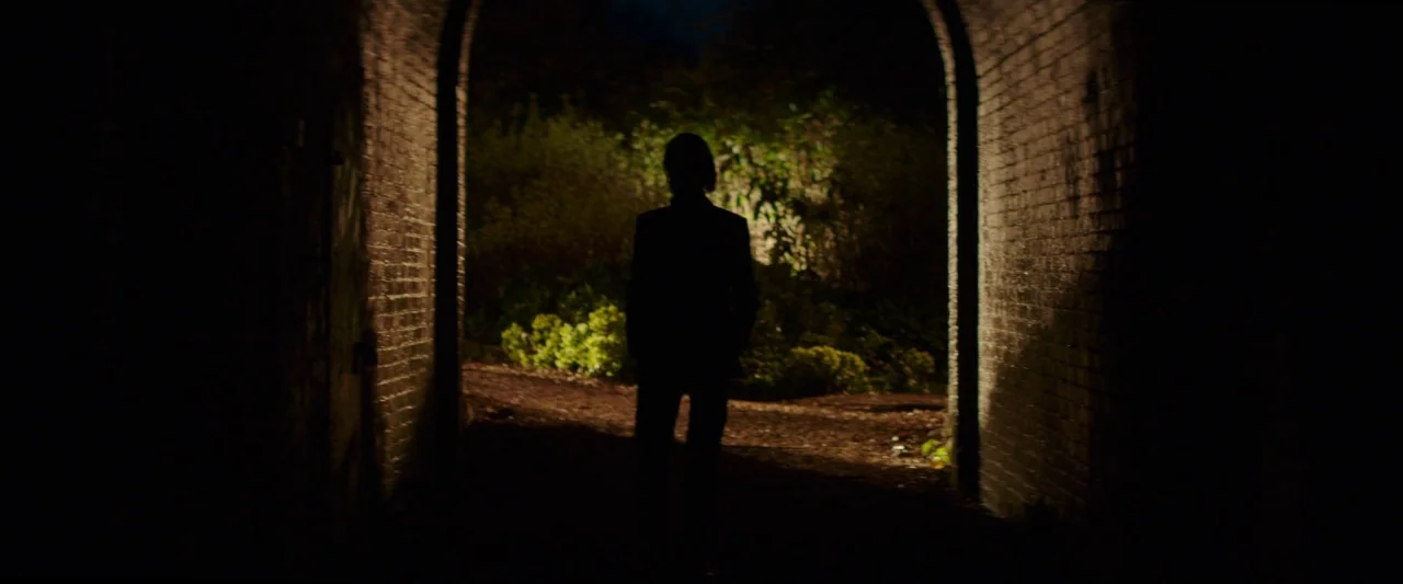 20,000 Days on Earth (2014) movie still: fullbody — A dark brick tunnel archway opens up to a patch of illuminated garden plants. A lone man stands…