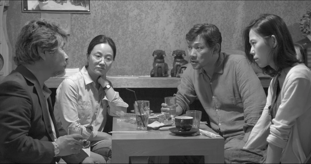 Grass (2018) movie still: medium — Four adults are seated at a wooden table in a dimly lit restaurant space. They are talking to each other while…