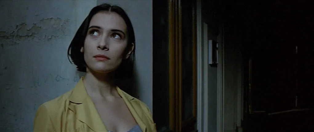 IP5 (1992) movie still: medium — A dimly lit narrow passage with a textured wall and dark shadowed doorways. A woman with short dark hair stands by…