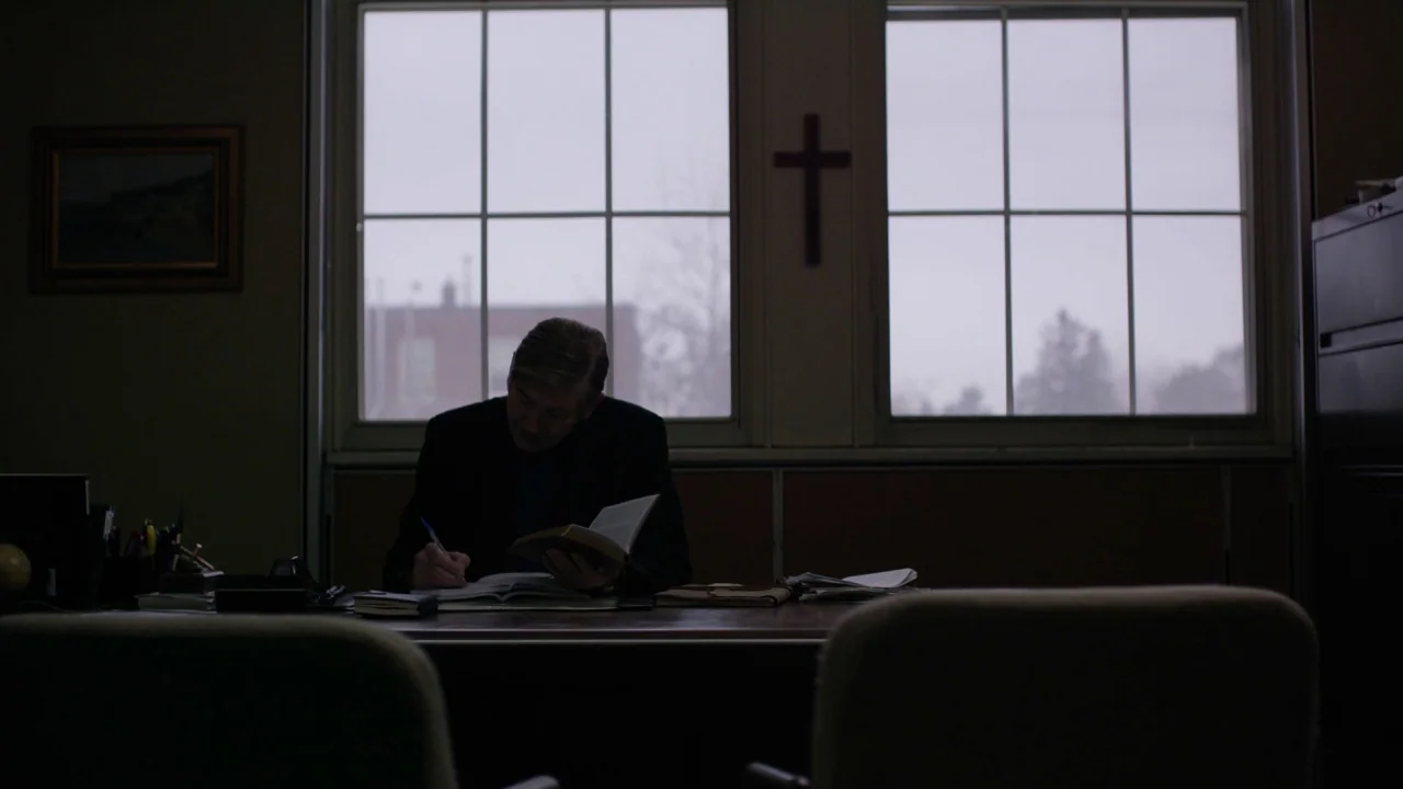 The Blackcoat’s Daughter (2015) movie still: medium — A dimly lit office features a wooden desk and windows overlooking a gray exterior. A man sits…