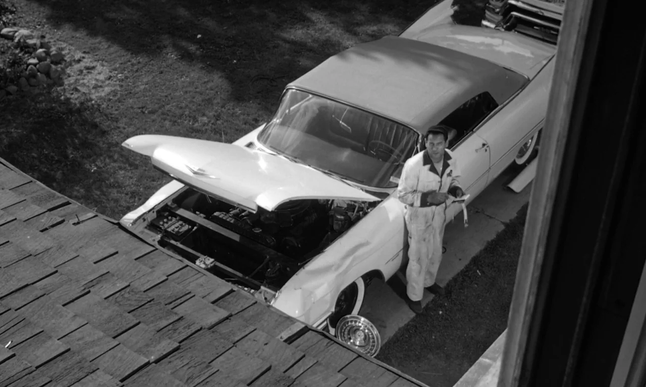 The Misfits (1961) movie still: wide — A man stands beside a white convertible with its hood open in a driveway. The vehicle has visible collision…