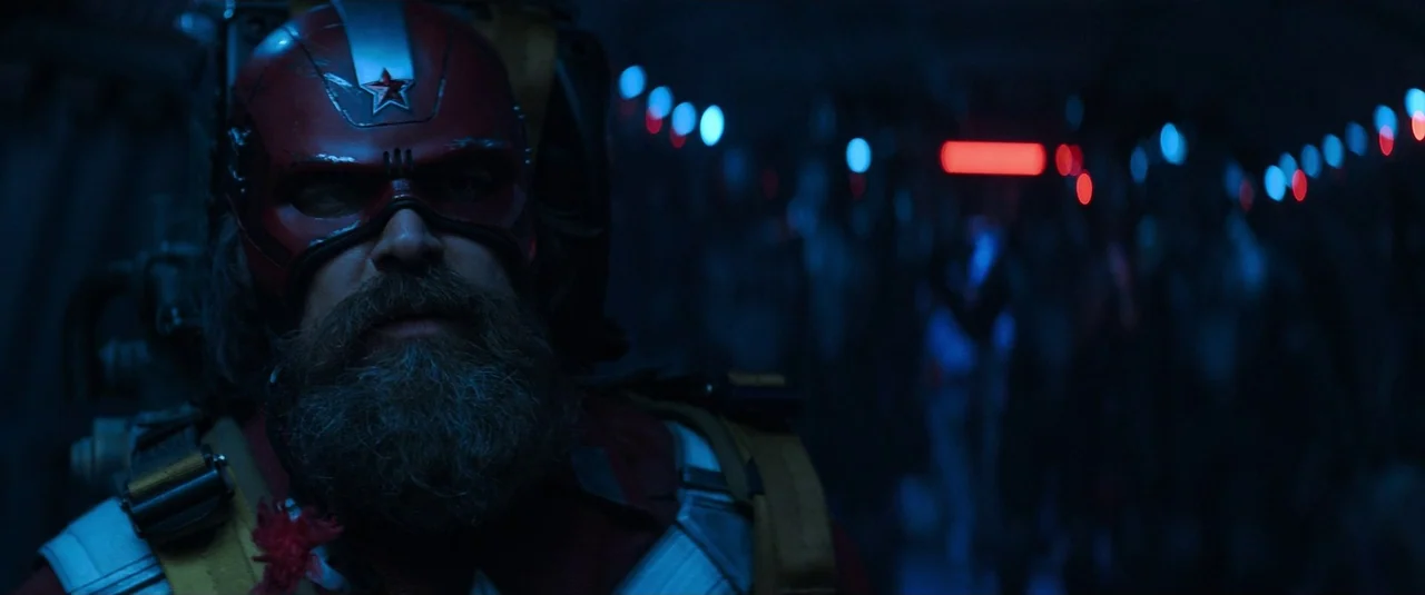 Black Widow (2021) movie still: closeup — A dimly lit aircraft cabin interior glows with faint blue and red emergency indicators. A bearded man in…