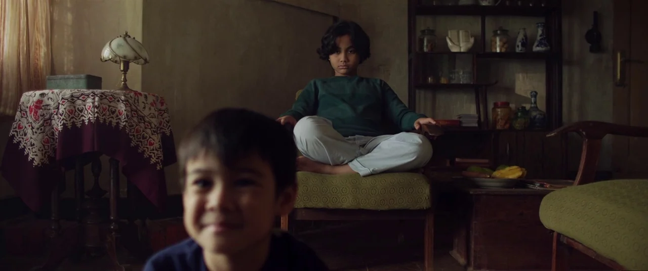 Satan’s Slaves (2017) movie still: medium — A cluttered room features a wooden shelf with jars and antique-style home decor. A young child in a…