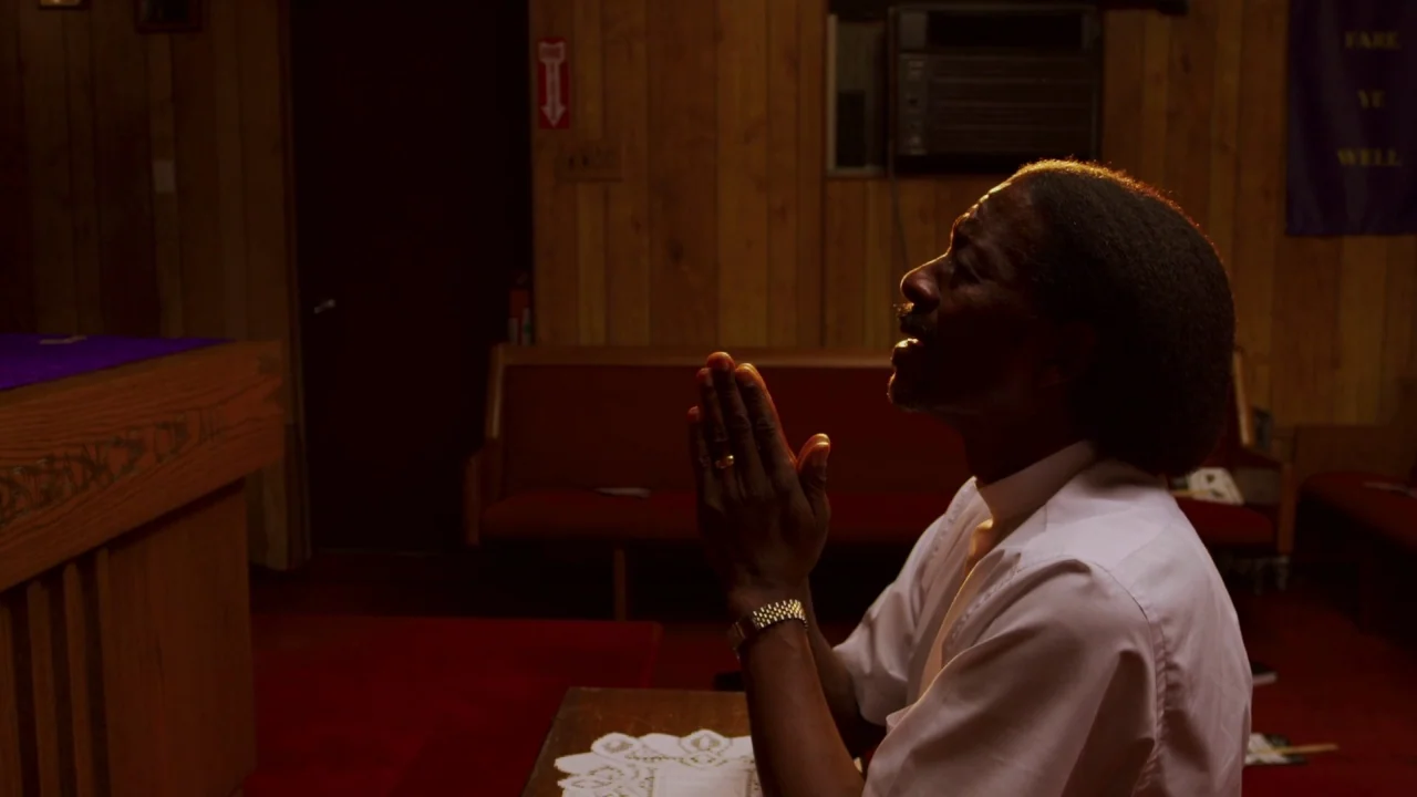 Red Hook Summer (2012) movie still: medium — A small, dimly lit room features wood-paneled walls and a wooden altar. An elderly man in a white…