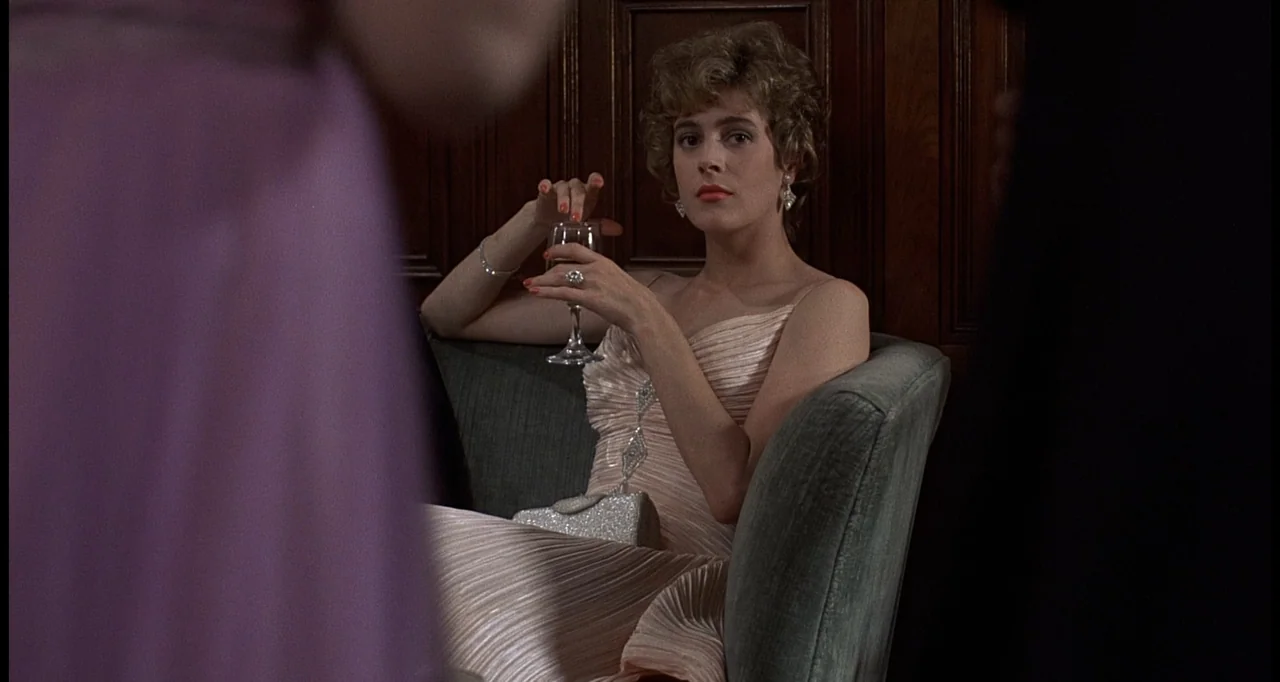 No Way Out (1987) movie still: medium — A wood-paneled room with a green velvet sofa and dark interior trim. A woman in an evening dress sits on the…