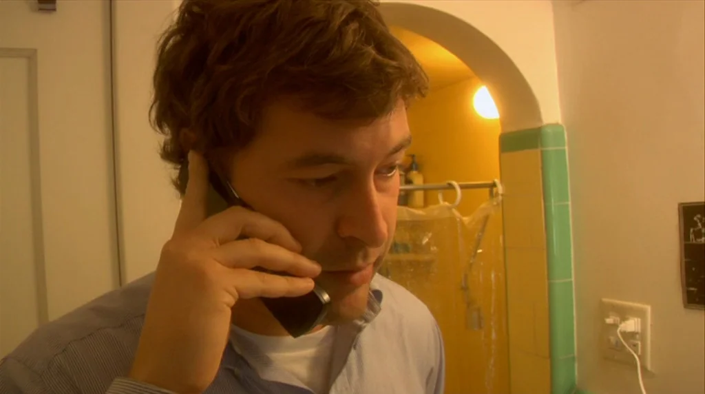 Humpday (2009) movie still: closeup — A man stands inside a bathroom with yellow and green tiled architectural features. He holds a black mobile…