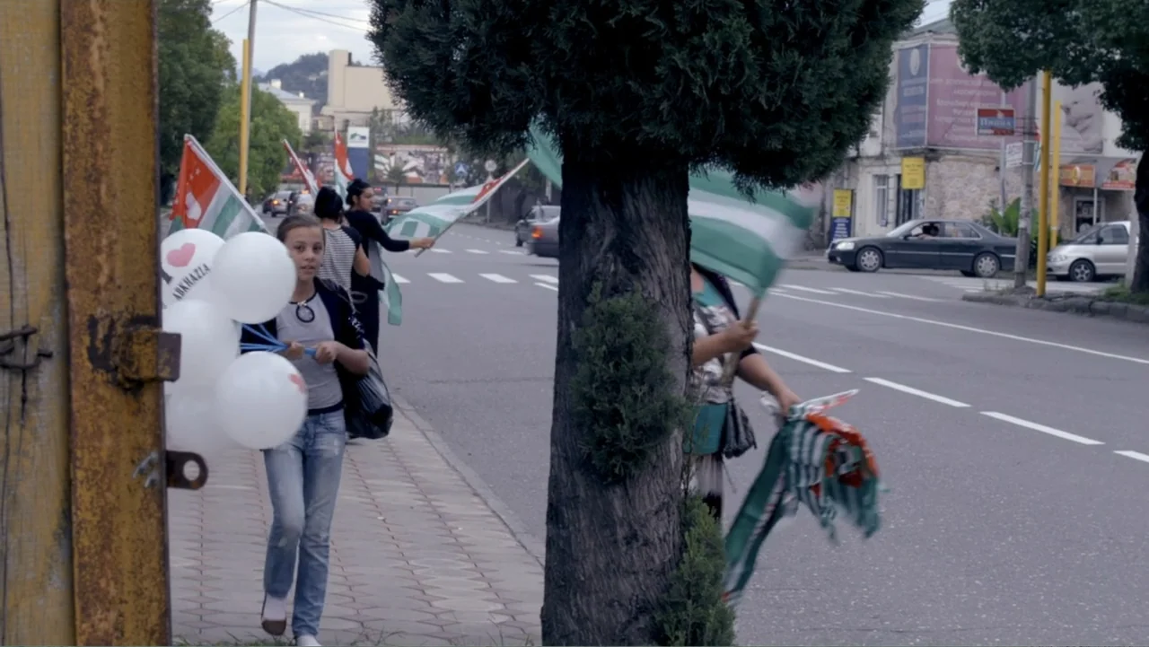 Letters to Max (2014) movie still: medium — A paved sidewalk runs alongside a paved multi-lane road near a roadside tree. A teenage girl holds…