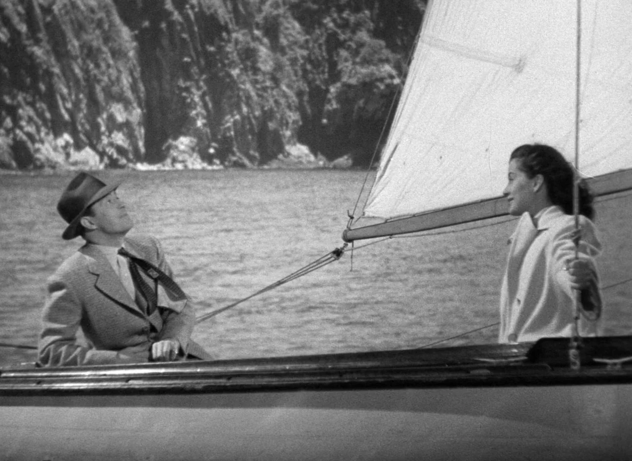 The Uninvited (1944) movie still: medium — A man and a woman sit aboard a small sailboat on the water. A rocky cliffside is visible in the background.
