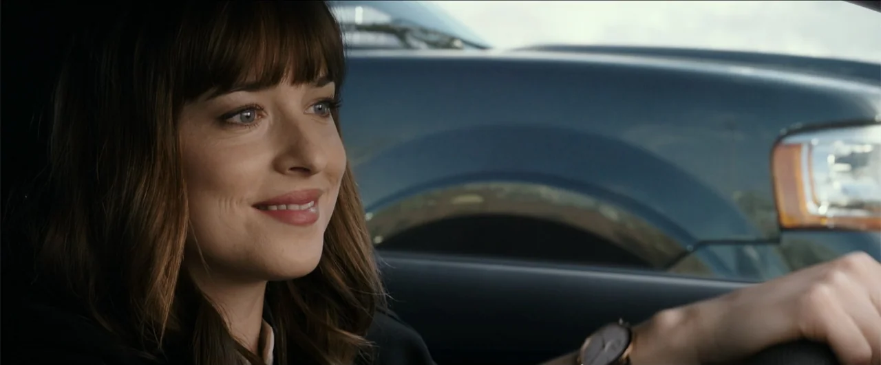 Fifty Shades Freed (2018) movie still: closeup — A woman sits in the driver's seat of a car during daylight. She maintains a slight smile as she…