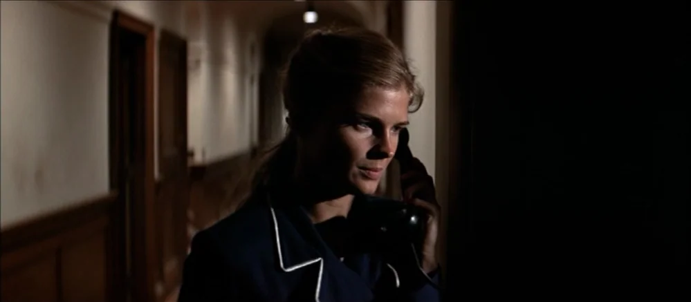 Carnal Knowledge (1971) movie still: medium — A dark, dimly lit corridor stretches back with closed wooden doors lining the walls. A woman in a blue…