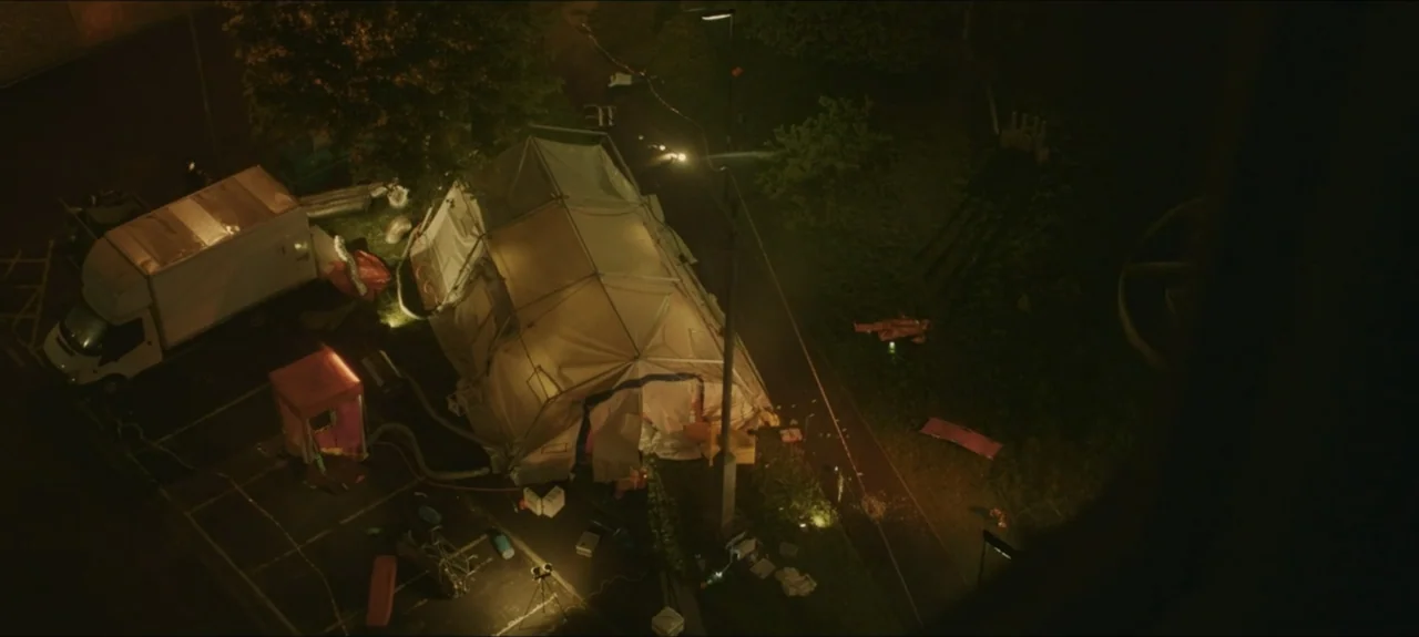 Containment (2015) movie still: wide — A top-down view shows an asphalt parking lot featuring a large portable white tent. A service truck is parked…