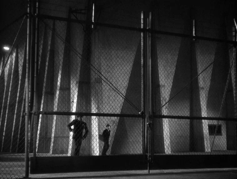 The Roaring Twenties (1939) movie still: wide — A dark night exterior features two men passing a high chain-link fence. The wall behind them is…