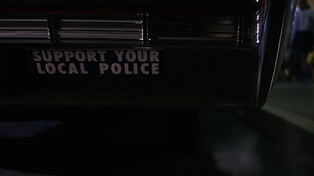 Bullitt (1968) movie still: closeup — The rear bumper of a dark vintage automobile features a rectangular political bumper sticker. The sticker…