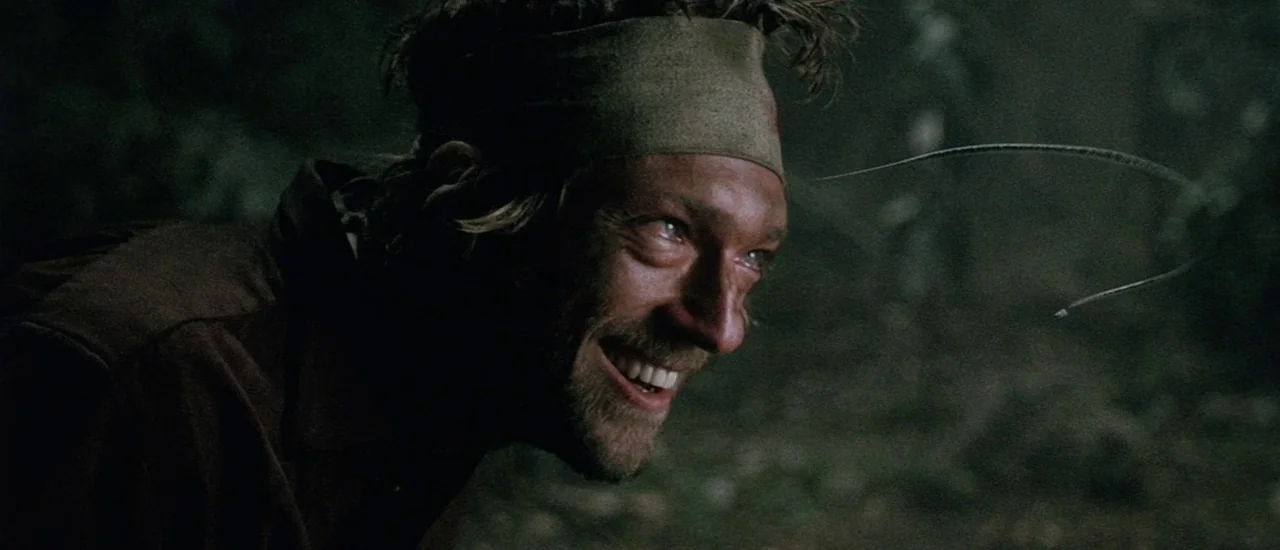 Renegade (2004) movie still: closeup — A person is in a dark, wooded area at night. A man with a headband looks to the side with an intense smile.