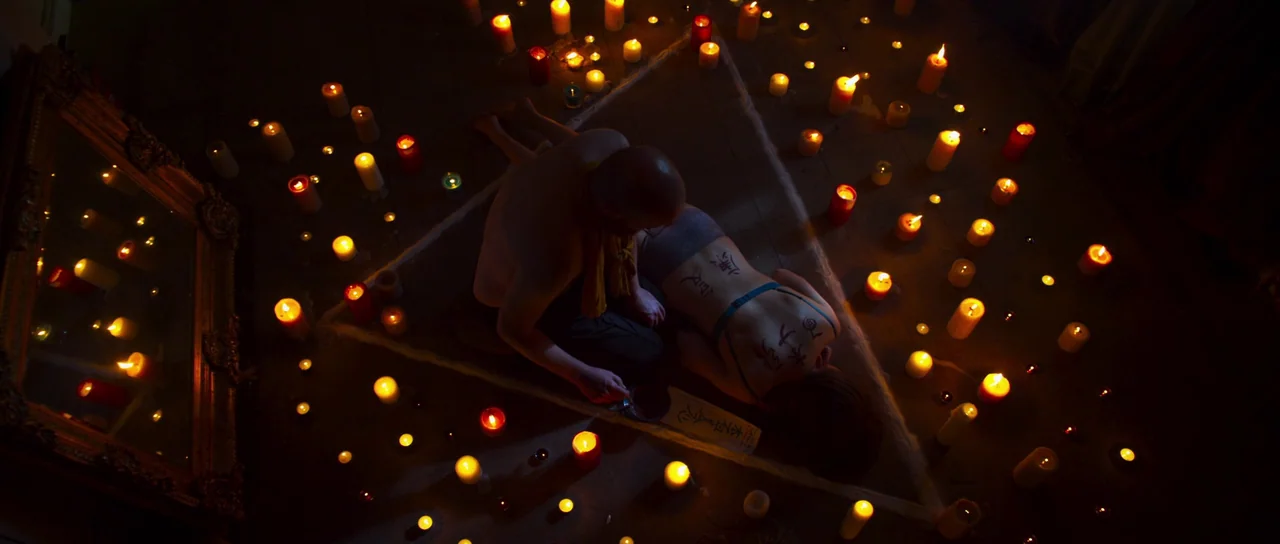 A Dark Song (2016) movie still: wide — A dark indoor space contains a large white triangle outlined on the floor. A man and a woman are positioned…