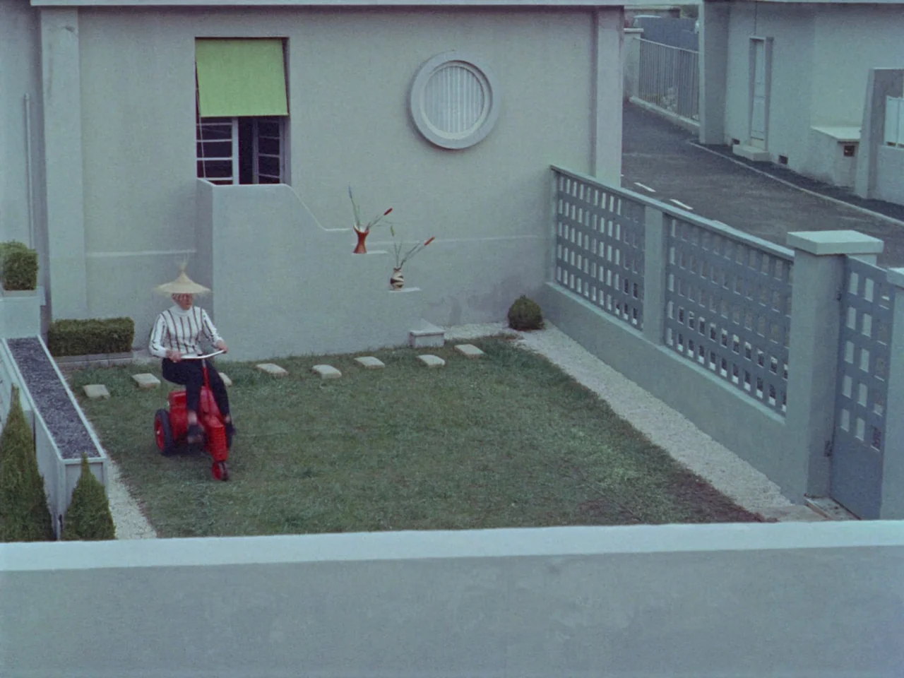 Mon Oncle (1958) movie still: wide — A minimalist modern house features a geometric courtyard with grey walls and a lawn. A woman wearing a conical…