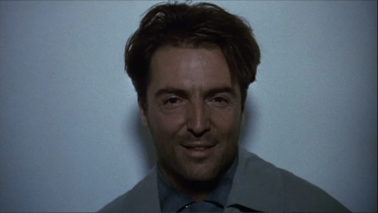 The Mambo Kings (1992) movie still: closeup — A man stands centered against a blank wall inside a dimly lit room. He maintains a frontal orientation…