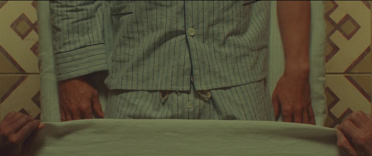Poison movie still: medium — A person lies under a sheet on a bed with patterned tiled edges visible. Hands enter the frame to pull the white sheet…