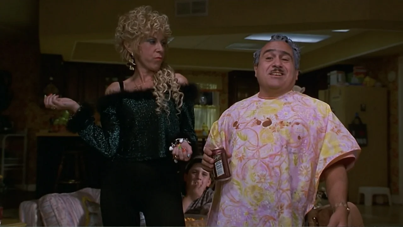 Matilda (1996) movie still: medium — A living room interior featuring wallpapered walls and generic home furniture. A woman and a man converse while…