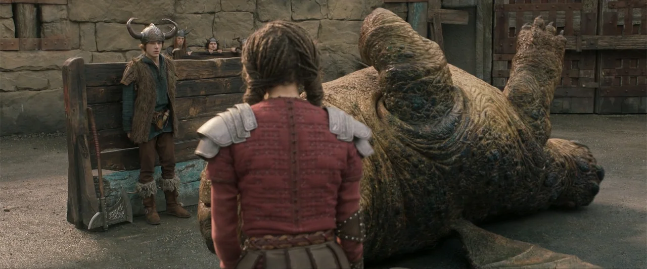 How to Train Your Dragon (2025) movie still: medium — An outdoor training arena features a stone wall and a large wooden gate. A woman faces a giant…