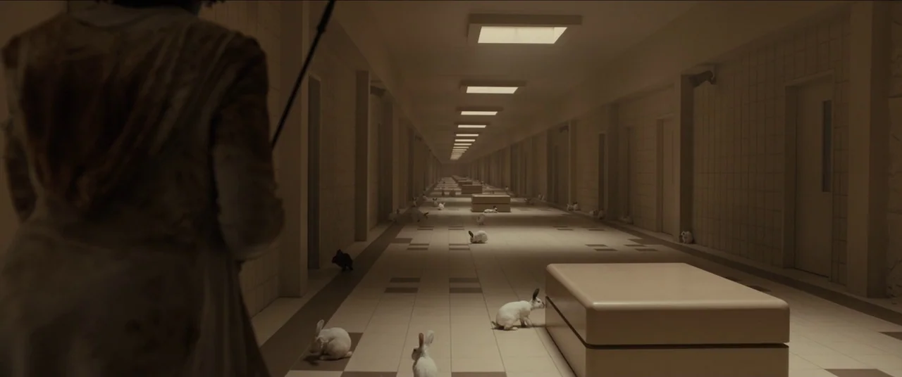 Us (2019) movie still: medium — A long, sterile tiled hospital corridor with repetitive doors and recessed square ceiling lights. A person stands in…